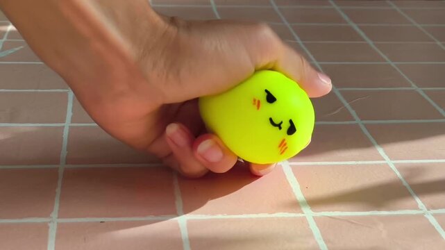 Hand squeezing a bright yellow squishy toy shaped like a dumpling, showing progressive deformation and playful interaction on a tiled surface with sunlight