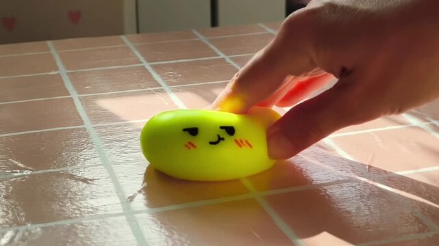 Hand gently presses and rolls a bright yellow squishy toy with a smiling face on a glossy tiled surface, showcasing playful interaction and tactile engagement