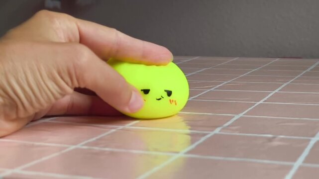Hand gently rolls a bright yellow squishy toy with a smiling face across a shiny pink tiled surface, showcasing the playful interaction and tactile quality of the object