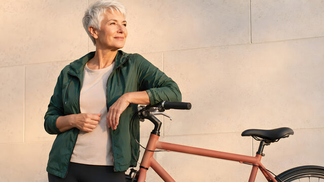 Older woman leaning on an orange bicycle against a pale stone wall, posing outdoors in casual clothing with a clean minimalist background.