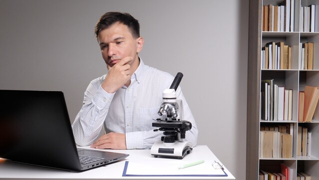 young man in a shirt sitting at a desk, contemplating a problem while looking at a laptop, with a microscope and books suggesting scientific research and education