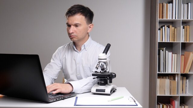 man focusing on his laptop, conducting scientific research and analysis in a laboratory or office setting with a microscope and bookshelf present