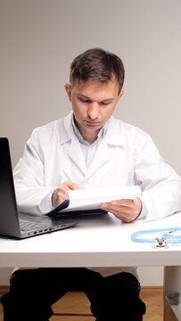 young male doctor in a lab coat at a desk using a laptop and notebook to document patient records, write prescriptions and manage telemedicine consultations and treatment plans