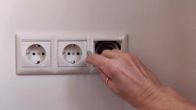 A hand inserts a black electrical plug into a wall outlet, household electrical use in a modern home interior, power and everyday safety.