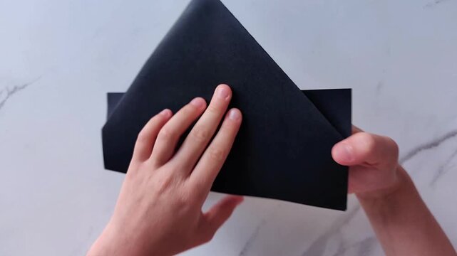 Two hands fold a black sheet of paper into a geometric shape, origami and creative paper craft on a light marble surface.