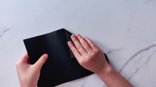 Two childs hands fold a black sheet of paper into a triangular shape on a marble surface, paper craft and creative handmade activity.