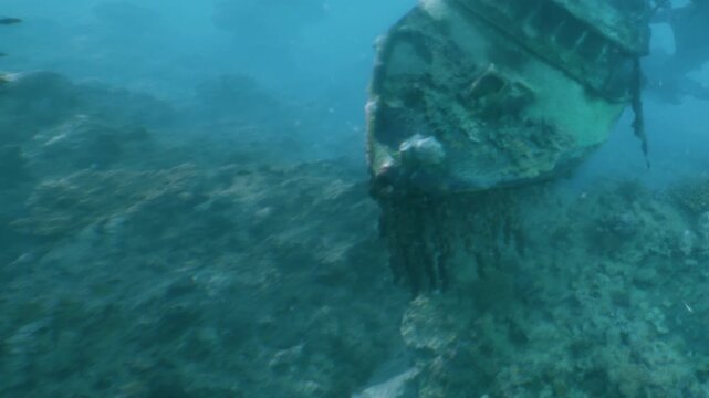 A sunken boat lies on the sea floor near a coral reef. The water is clear, allowing a view of the marine life around the site. Small fish swim nearby as sunlight filters through the water.