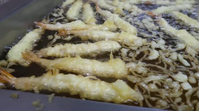 High-angle close-up video footage of multiple battered prawns frying inside a commercial deep fryer. The Japanese-style ebi tempura pieces bubble rapidly in hot, golden boiling oil as they cook to a p