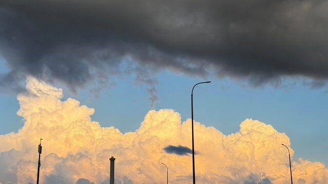 Massive golden cumulus clouds illuminated by setting sun under a heavy dark storm cloud canopy, with silhouettes of city streetlights and a tower.