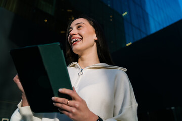 Cheerful young woman with braces laughing while holding digital tablet outdoors in city. Happy girl enjoying success or positive news on mobile device in front of modern office building at sunset. © BullRun