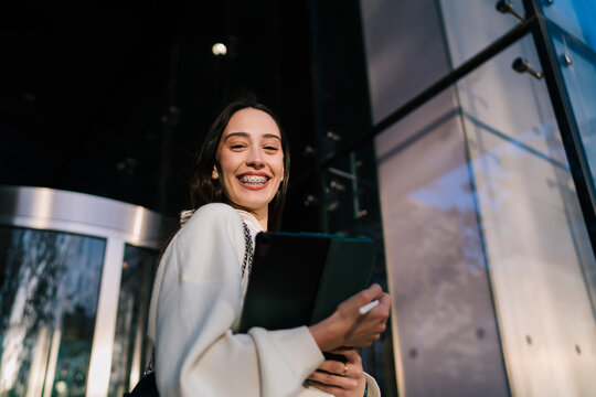 Portrait of happy young woman with braces laughing while holding digital tablet outdoors. Cheerful female model near modern glass building, social media influencer and city connectivity concept.