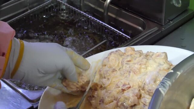 A commercial kitchen setting showcasing the process of deep frying battered chicken. A chef is seen using metal baskets to submerge food into hot, bubbling oil, while a white plate in the foreground h