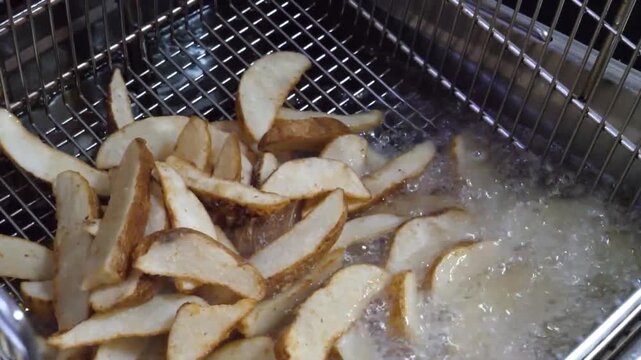 Close-up shot of thick-cut, skin-on potato wedges being lifted out of bubbling hot oil in a stainless steel deep fryer basket.