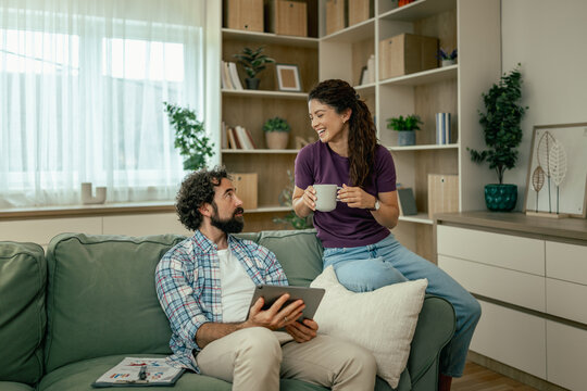 Couple relaxing at home on sofa chatting over coffee while using a tablet in living room