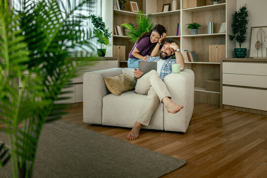 Couple relaxing at home in cozy living room using tablet amidst indoor plants and bookshelves