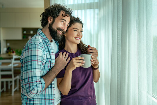 Young couple enjoying morning coffee together at home looking out window with warm smiles