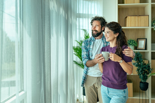 Young couple standing by window smiling and enjoying coffee together in bright living room