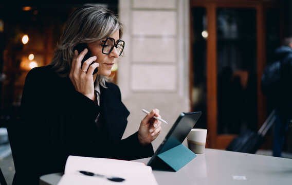 Caucasian businesswoman in stylish glasses talking on mobile phone at terrace table, middle aged female professional using stylus and tablet for work, remote office and digital lifestyle.