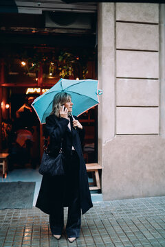Full length of elegant mature woman holding umbrella and talking on phone at city street, female professional in black coat having business conversation in rain, urban lifestyle concept