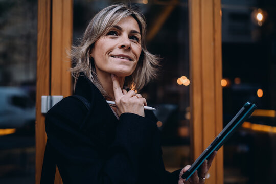 Smiling mature woman holding digital tablet and stylus pen looking away, confident female entrepreneur with positive attitude, creative professional in urban setting, authentic representation.