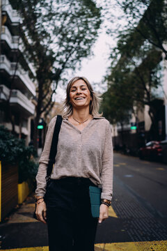 Joyful mature female professional laughing on urban street holding tablet, happy woman enjoying business success and digital nomad life, authentic emotional state of creative entrepreneur in city.