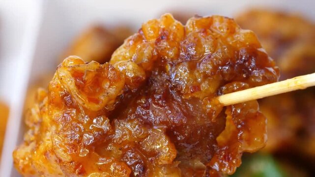 A macro slow-motion shot focusing on a single piece of double-fried Korean chicken glazed in a sticky, glossy gochujang sweet chili sauce. The juicy, deep-fried chicken nugget is lifted with a wooden 