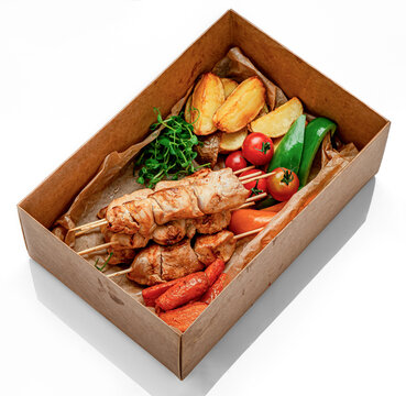 Grilled chicken skewers with potatoes and tomatoes in box. Chicken shish kebabs are packed with roasted potato wedges, carrots, bell peppers, and cherry tomatoes in a brown card box.