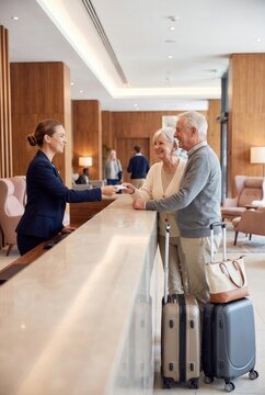 Receptionist assisting elderly couple with check-in at a modern hotel lobby featuring wooden accents and comfortable seating areas with luggage nearby