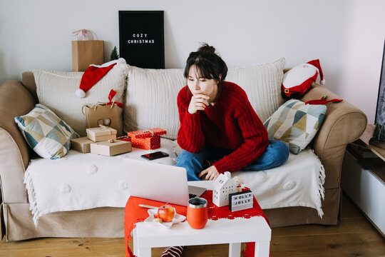 Woman in knitted sweater works on laptop on sofa beside wrapped parcels and HOLLY JOLLY sign. Remote work december, laptop home office christmas, finishing tasks before holidays