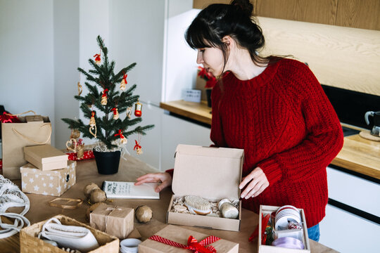 Woman holds notebook while checking open box with brush and jar in kitchen. Small business checking order list, verifying christmas order contents, matching gift to checklist