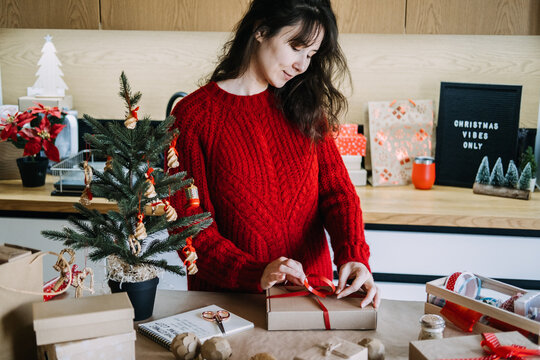 Woman hands hold kraft box tied with ribbon above table with wrapping supplies. Mindful gift giving concept, quality over quantity christmas, intentional consumerism holiday