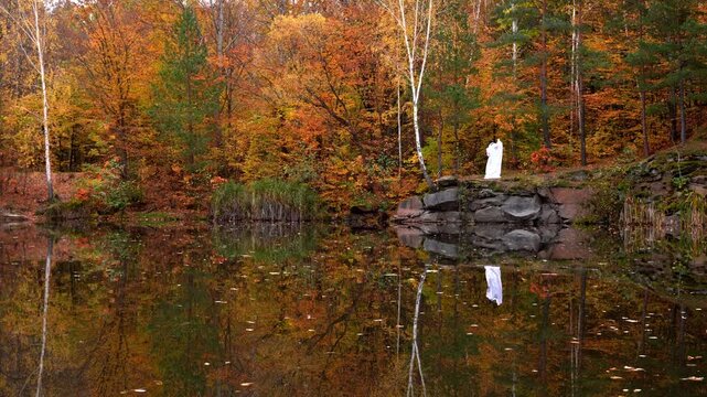 Playful ghost costume made from white bedsheet stands in autumn forest reflection on water lake surface, wearing glasses and cap. The humorous character creates festive seasonal atmosphere inspired by