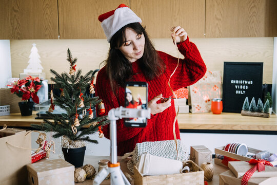 Woman in Santa hat cuts raffia ribbon before phone on tripod near CHRISTMAS VIBES ONLY sign. Handmade christmas gift wrapping business, christmas craft supplies small shop, artisan christmas wrapping 