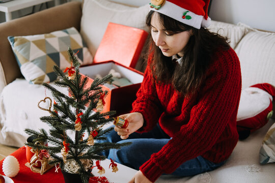 Young woman in Santa hat hangs small ornament on miniature Christmas tree while sitting on sofa. Small space holiday decorating, handmade straw ornaments, minimalist christmas, slow living traditions