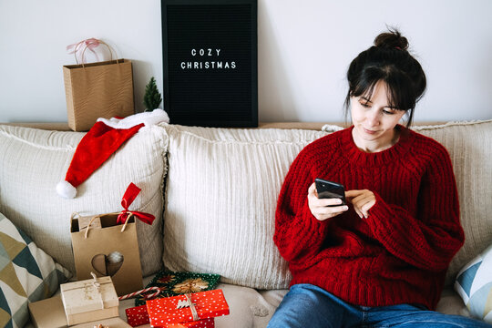 Woman uses smartphone on sofa beside wrapped gifts and COZY CHRISTMAS sign. Sending christmas wishes phone, holiday greeting message, texting family december