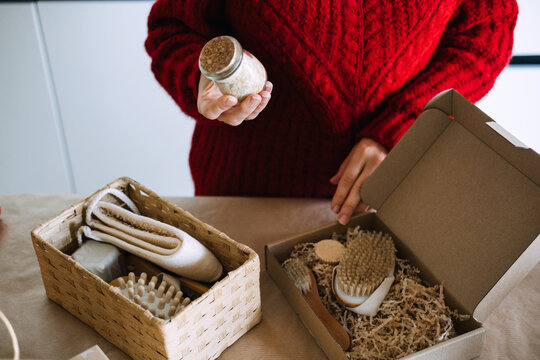 Woman holds jar of bath salt above open kraft box with brushes and paper filler. Quality control handmade products, checking product before shipping, artisan quality assurance process