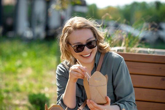 Joyful woman holds Asian takeaway food for summer lunch break. Happy person enjoys tasty noodles on city street during sunny day.