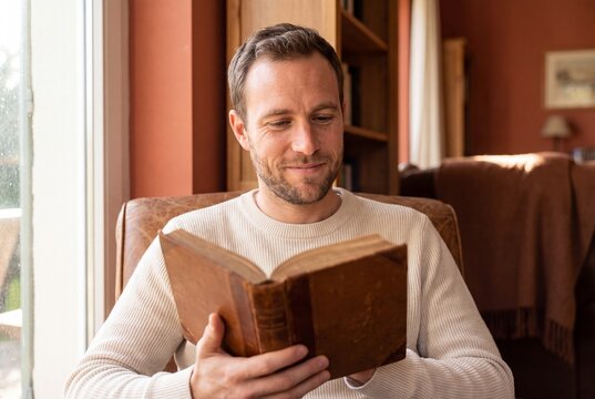Smiling Man Reading Antique Leather Book Sitting in Armchair by Sunlit Window