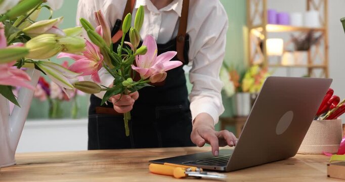 Florist using laptop while making bouquet of beautiful lilies at wooden table in shop, closeup