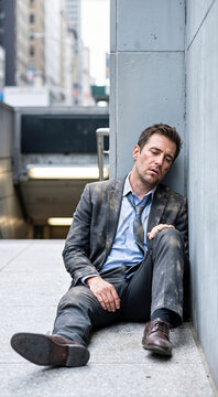 Exhausted businessman in dusty charcoal suit leaning against concrete wall outdoors on urban city street. Tired stressed male executive feeling defeated after work failure vertical shot