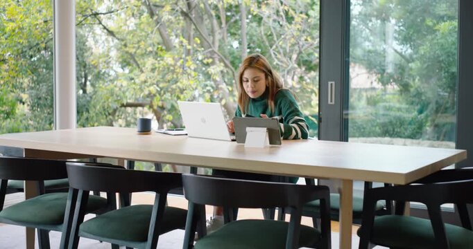 African American woman sitting at dining table, using laptop and tablet, taking phone call for work