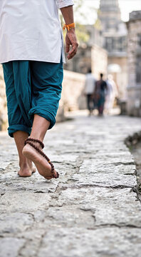 Barefoot male pilgrim walking on an ancient stone pathway wearing a traditional rudraksha bead foot ornament. Rear view close-up of feet on cobblestone street in India near temple background