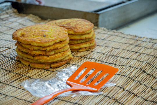 Freshly cooked mung bean pancakes bindaetteok stacked on bamboo mat with orange spatula at traditional local street market in South Korea.
