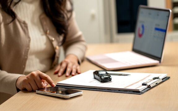 Woman using smartphone calculator with car model on clipboard beside laptop displaying financial dashboard