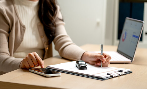 Woman calculating car loan with smartphone and miniature car model on clipboard beside laptop