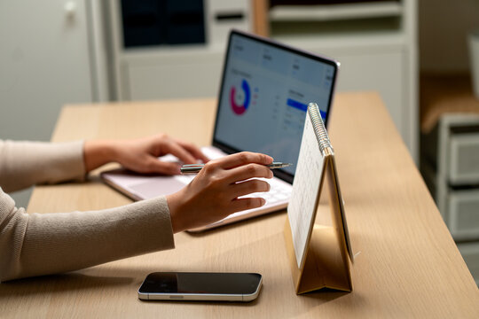 Person reviewing financial dashboard on laptop with pen poised beside standing desk calendar