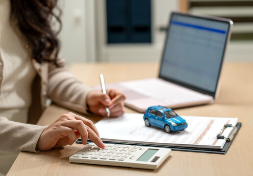 Woman calculating car loan with blue model vehicle on clipboard beside laptop
