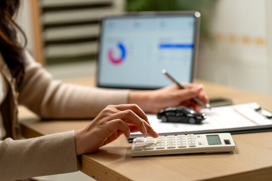 Woman calculating car loan with calculator and miniature vehicle on clipboard beside laptop