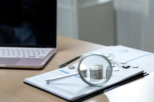 Magnifying glass focusing on coin stacks atop financial charts beside laptop on wooden desk