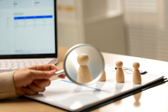 Hand holding magnifying glass over wooden person figurines on financial documents beside laptop